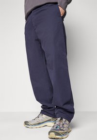 Homme portant un pantalon décontracté bleu marine et des chaussures de randonnée grises et beiges, se tenant avec une main dans sa poche.