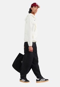 Biała kurtka z kapturem, czarne luźne spodnie, czarne sneakersy z białymi paskami oraz bordowa czapka. Gładki materiał z minimalnym brandingiem i akcentami.