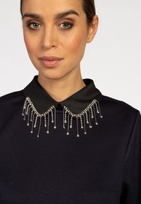 Schwarzes Oberteil mit einem strukturierten Kragen, der mit silbernen Perlen und herabhängenden Kristallakzenten verziert ist. Glatte Stoffoberfläche. Elegantes Detail.