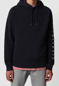 Felpa con cappuccio blu navy a maniche lunghe. Presenta un testo grafico bianco lungo la manica e un orlo a costine. Indossata sopra una maglietta rosa e pantaloni della tuta grigi.