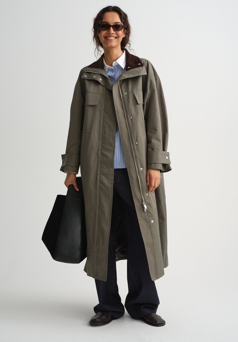 Femme portant un long manteau vert olive, chemise rayée, pantalon foncé, lunettes de soleil, tenant un grand sac noir, souriante devant un fond uni.