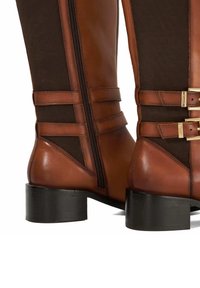 Bruine leren kniehoge laarzen met een textielpaneel, rits aan de zijkant en gouden hardwareaccenten op de banddetails. Blokhak.