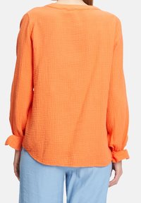 Camicia arancione a maniche lunghe, con una texture e un collo rotondo. Presenta polsini arricciati e un orlo leggermente curvato, realizzata in tessuto leggero.