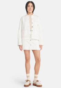 Hvit denimjakke med to store lapperlommer, sammen med et hvitt skjørt. Under er en stripete topp. Brune platåsko og stripete sokker.