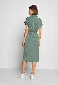 Abito verde a maniche corte con colletto, vita aderente e lunghezza al ginocchio. Tessuto liscio e spacchi laterali, abbinato a sneakers bianche.