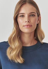 Blond vlasy s vlnami, oblečená v námorníckom modrom topi s okrúhlym výstrihom. Pokožka je hladká s prirodzeným makeupom; oči sú zelené.