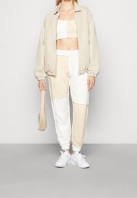 Bežová oversized bunda, bílé a béžové kalhoty s barevnými bloky, krátký béžový top a bílé tenisky. Součástí je malá béžová taška.