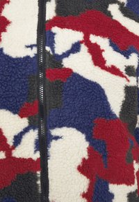 Fleecejacka med dragkedja fram, med ett kamouflage-mönster i röd, vit, grå och blå. Den texturerade ytan kontrasterar med den släta dragkedjan.