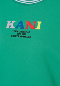 Tessuto verde con lettere colorate "KANI" e il testo "THE ORIGINAL EST. 1989 REG. TRADEMARK" stampato in nero sotto.