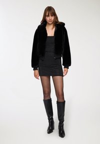 Jacket bomber in pelliccia sintetica nera con colletto oversize, indossato sopra un vestito attillato nero a pois e stivali neri con tacco al ginocchio.