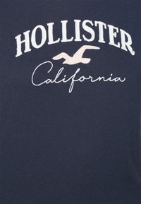 Tissu bleu marine avec le texte brodé en blanc "HOLLISTER Californie" et un petit logo de mouette rose entre les mots.
