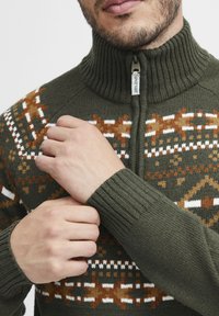 Pulover verde tricotat cu gât înalt, dotat cu fermoar, modele geometrice în nuanțe de brun, alb și portocaliu, și manșete ribbate pentru o fixare comodă.