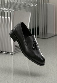 Mocasines de cuero negro con punta redondeada, textura suave y borlas decorativas en negro. Diseño minimalista sin acentos de hardware visibles.
