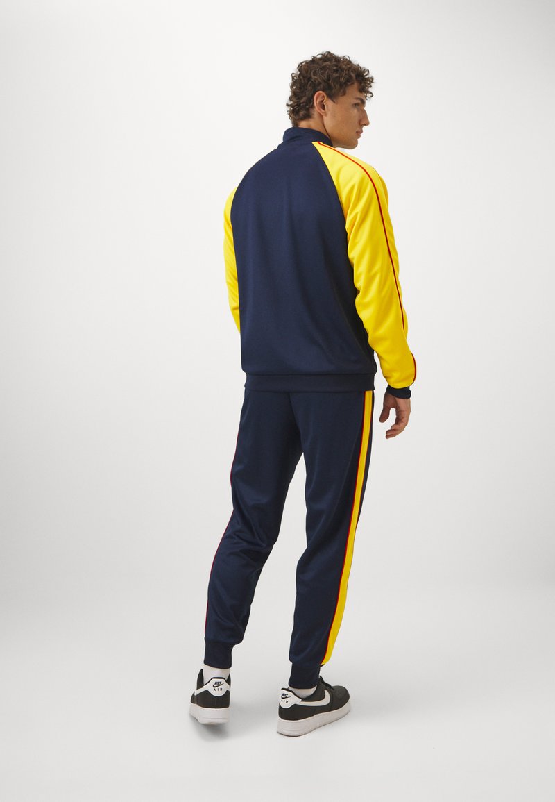 Tuta navy e gialla con maniche lunghe, dettagli a righe a contrasto e pantaloni aderenti; abbinata a sneaker bianche e nere.