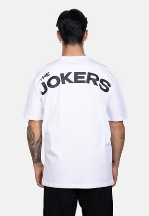 Persona con cabello corto y oscuro lleva una camiseta blanca con texto negro "THE JOKERS" en la parte superior de la espalda sobre un fondo liso.