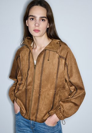 Jonge vrouw met lang bruin haar, gekleed in een bruine geperforeerde ritsvest en een blauwe spijkerbroek, staand met handen in de zakken tegen een effen muur.