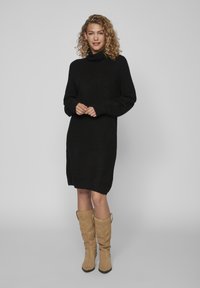 Donna con capelli ricci che indossa un vestito maglione a collo alto nero e stivali alti fino al ginocchio in camoscio beige, in piedi su uno sfondo neutro.