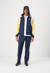 Veste bleu royal avec des manches jaunes, fermeture éclair et un logo. Pantalons bleu royal assortis avec des accents jaunes, portés avec des chaussures de sport noires.