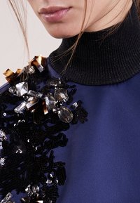 Marinblus med en ribbad svart polokrage; dekorerad med paljetter och ädelstenar i svarta, guldiga och klara material.