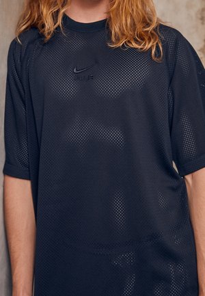 Personne aux cheveux longs portant un t-shirt noir perforé Nike Air à manches courtes, debout devant un fond neutre.