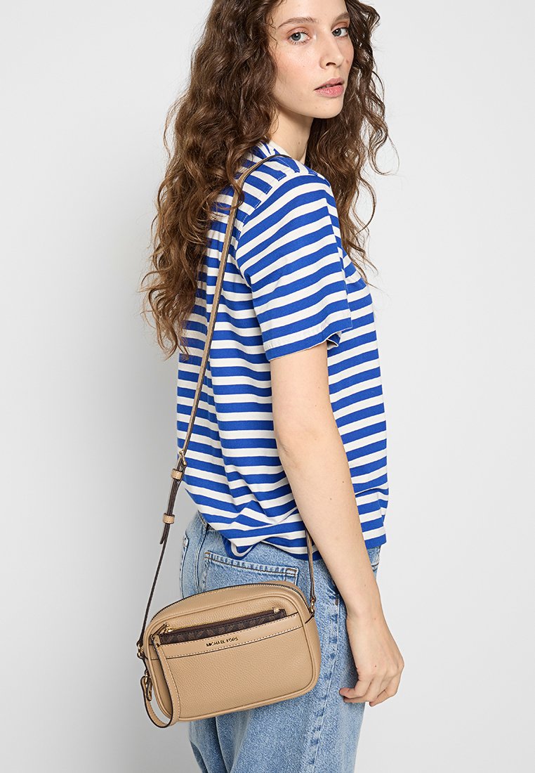 Bolso crossbody beige con un acabado texturizado, que cuenta con un bolsillo frontal con cremallera y una correa ajustable, llevado sobre una camisa a rayas.