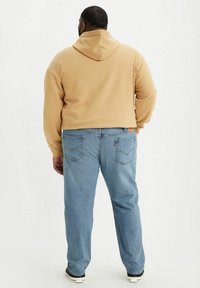 Tan hoodie with a hood, ribbed cuffs, paired with light blue jeans featuring back pockets and a woven label. Casual footwear completes the look.