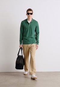 Cămașă polo verde cu mânecă lungă, pantaloni bej, purtând o geantă sport neagră. Tenisi albi, ochelari de soare negri. Design minimalist, croială lejeră.
