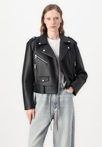 Black faux leather cropped motorcycle jacket with silver hardware, zippers, and a pointed collar; worn over a white t-shirt and light denim jeans.