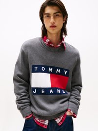 Pulover tricotat gri cu o bandă proeminentă albastră, albă și roșie ce scrie "TOMMY JEANS." Purtați peste o cămașă în carouri roșii și negre.