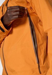 Oranje jack met een gladde textuur, voorzien van een ritszak. De rits is bruin, wat contrasteert met de stof. Er is accentstiksel zichtbaar.