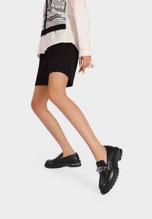 Scarpe in pelle nera con suola spessa, caratterizzate da un dettaglio con fibbia argentata e una superficie liscia, abbinate a pantaloni corti neri e una camicia bianca.