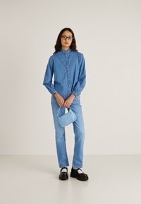 Denimskjorte med knappestengning, høy krage og lange ermer, kombinert med lyseblå jeans og svarte platåsko. Holder en liten blå håndveske.