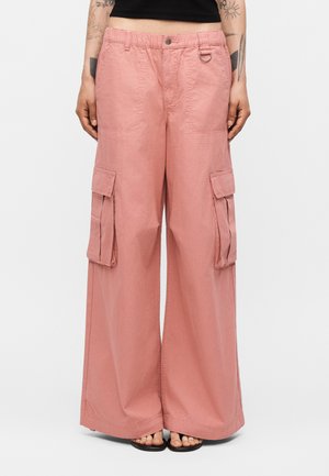 Άτομο που φορά φαρδιά ροζ παντελόνια cargo με μεγάλες πλευρικές τσέπες, ελαστική μέση και μαύρο κοντό τοπ, στέκεται ξυπόλυτο σε σανδάλια.