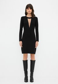 Schwarzes Langarmkleid mit tiefem V-Ausschnitt, taillierter Form und glattem Stoff. Kombiniert mit knielangen schwarzen Stiefeln, die elegante Details hinzufügen.