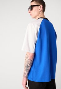 Blue and grey color-block T-shirt with a relaxed fit, short sleeves, and a smooth texture. Features a black neckline and visible tattoos on the arm.