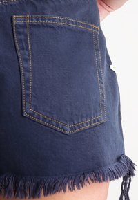 Pantalones cortos de mezclilla azul oscuro con un dobladillo deshilachado, que cuentan con un bolsillo trasero y costuras amarillas a lo largo de las costuras para un detalle adicional.