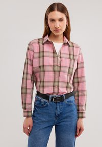 Camicia a quadri rosa e verde con colletto button-down, maniche lunghe e tasca sul petto, abbinata a jeans in denim blu e una cintura nera.