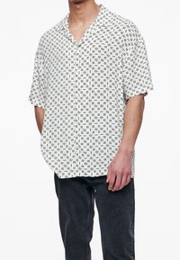 Camicia a maniche corte bianca con motivo geometrico nero. Presenta un colletto classico e una vestibilità ampia. Il materiale sembra leggero e liscio.