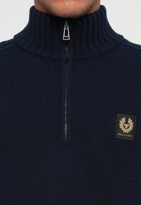 Pulover tricotat bleumarin cu guler înalt cu nervuri, fermoar frontal și un patch pătrat negru cu un design cu aripi pe partea stângă a pieptului.