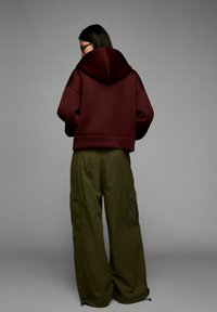 Person with long dark hair and glasses wearing a burgundy hooded sweatshirt and loose olive green cargo pants, facing away on a gray background.