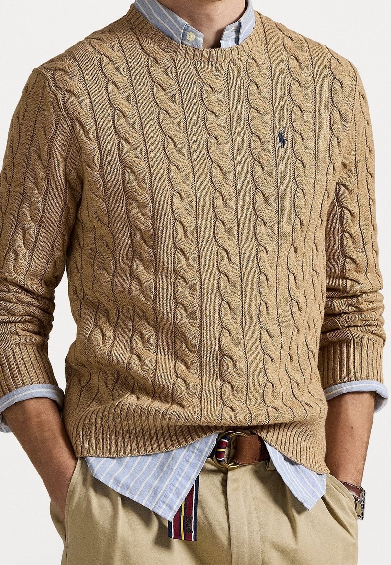 Homme portant un pull en maille torsadée beige par-dessus une chemise bleue rayée avec les manches retroussées, un pantalon beige et une ceinture rayée, les mains dans les poches.
