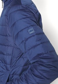 Marineblauwe pufferjas met gewatteerde textuur en geïsoleerd ontwerp; voorzien van een klein GAP-logo op de linkerschouder.