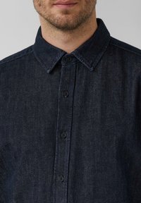 Camicia in denim scuro con abbottonatura frontale, colletto a punta e bottoni neri. Presenta un tessuto testurizzato con un delicato motivo a trama.