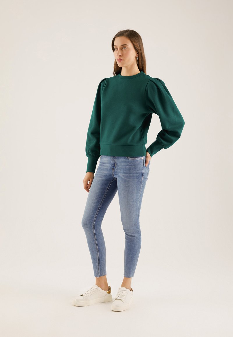 Green sweatshirt with puffed sleeves, ribbed cuffs, and hem. Paired with fitted blue jeans and white sneakers featuring a gold accent.
