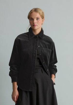 Jeune femme aux cheveux blonds portant une chemise ample en velours côtelé noir sur un pantalon sombre, debout devant un fond gris clair uni.