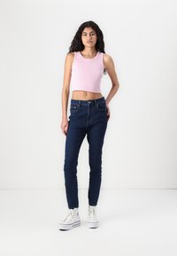 Roza rebrasta kratka majica u kombinaciji s tamnoplave skinny traperice viskog struka i bijelim sportskim tenisicama. Jednostavna pozadina, u stojećem stavu.