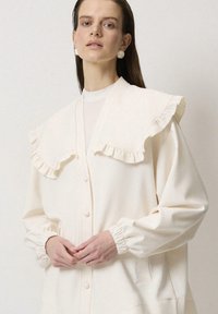 Chaqueta blanca roto tipo camisa con collar rizado, mangas fruncidas y bolsillos laterales. Fabricada en un tejido suave con una textura lisa.