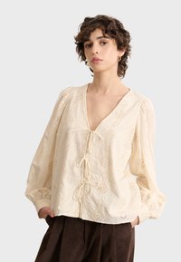 Cremfarbene Bluse mit tiefem V-Ausschnitt und Puffärmeln. Verfügt über gestickte Diamantmuster und ein vorderes Schnürdetail.