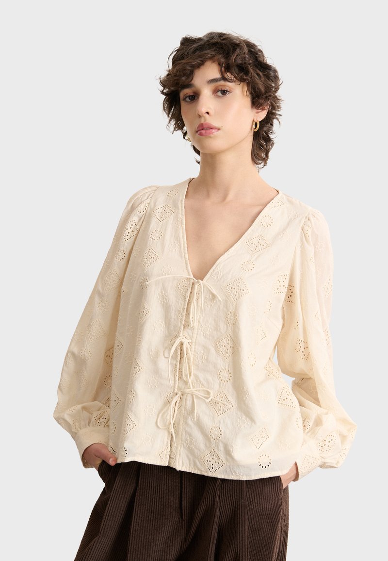 Cremfarbene Bluse mit tiefem V-Ausschnitt und Puffärmeln. Verfügt über gestickte Diamantmuster und ein vorderes Schnürdetail.