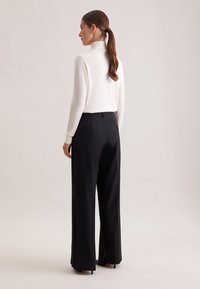 Biała sweter z małym golfem w zestawieniu z czarnymi spodniami o szerokiej nogawce, charakteryzującymi się gładką fakturą i dopasowanym krojem, stylizowany na wysokich obcasach.